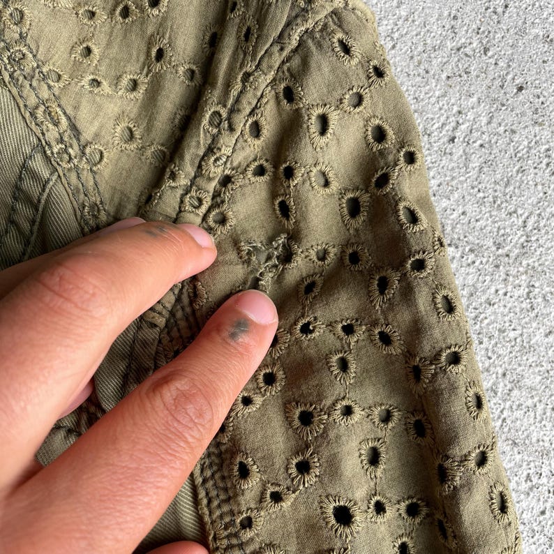 Pode incluir: Close-up de tecido verde-oliva com bordado de ilh&oacute;s intrincado. O tecido &eacute; texturizado e detalhado, com uma costura vis&iacute;vel e uma m&atilde;o tocando o material. O design de ilh&oacute;s cria um visual delicado e arejado.