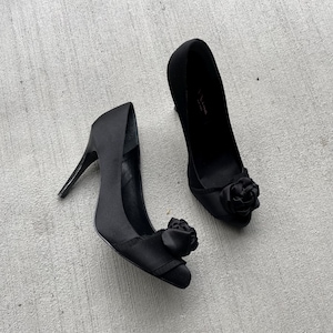 Op de afbeelding: Een paar zwarte satijnen pumps met een bloemenaccent op de neus. De ene schoen heeft een strik op de neus en de andere een roos.