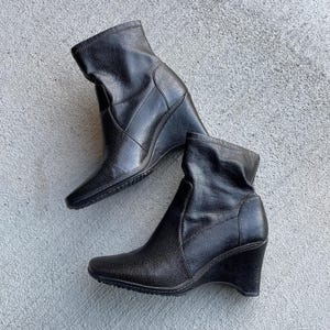 Könnte beinhalten: Ein Paar schwarze Stiefeletten mit Keilabsatz. Die Stiefel haben ein schlankes, modernes Design mit spitzer Zehenpartie und einer strukturierten Lederoptik. Der Keilabsatz bietet sowohl Stil als auch Halt. Die Stiefel stehen vor einem neutralen Hintergrund.