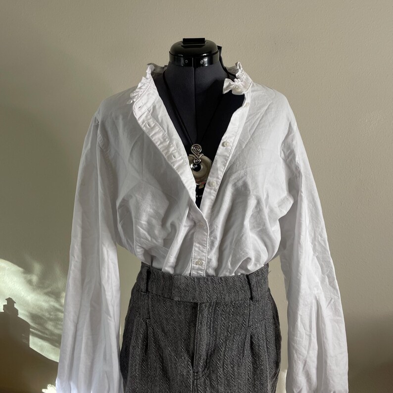 May include: A white button-down shirt with a ruffled collar and long sleeves is paired with gray herringbone trousers. A silver pendant necklace hangs over the shirt. The outfit is displayed on a black mannequin.