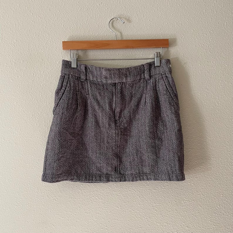 May include: A gray herringbone mini skirt hanging on a wooden hanger. The skirt has a classic design with a zipper and pockets. The fabric has a textured appearance.