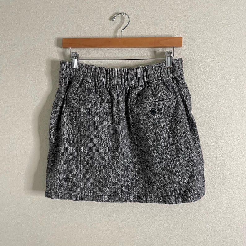 May include: A gray herringbone pattern skirt with an elastic waistband and two back pockets. The skirt is hanging on a wooden hanger against a neutral background. The skirt has two black buttons on the back pockets.