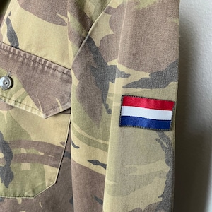 Może przedstawiać: Kurtka w kamuflażu z czerwoną, białą i niebieską naszywką na rękawie. Naszywka przedstawia flagę Holandii.
