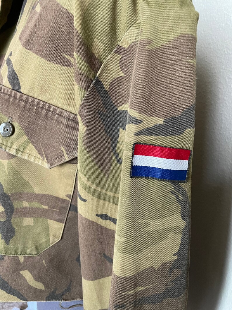 Może przedstawiać: Kurtka w kamuflażu z czerwoną, białą i niebieską naszywką na rękawie. Naszywka przedstawia flagę Holandii.