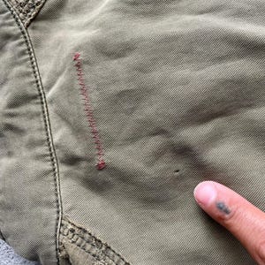 Pode incluir: Close-up de tecido verde-oliva com costuras vis&iacute;veis e um pequeno buraco. Uma se&ccedil;&atilde;o da costura da pe&ccedil;a &eacute; vis&iacute;vel, juntamente com uma linha de costura vermelha. Um dedo com uma tatuagem aponta para o tecido.