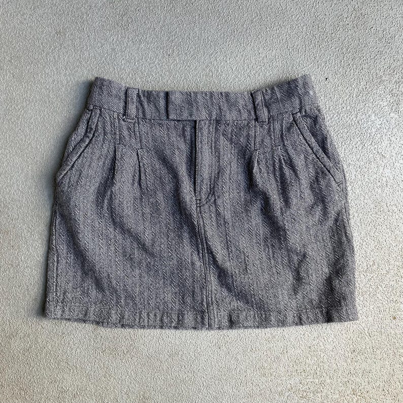 May include: A gray herringbone mini skirt with a waistband, belt loops, and front pockets. The skirt has a zipper and button closure. The fabric has a subtle vertical stripe pattern.
