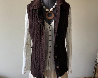 Könnte beinhalten: Eine dunkelbraune Strickweste mit Zopfmuster, Kragen und Knopfverschluss. Die Weste ist über einer weißen Bluse und einem cremefarbenen Rock getragen. Eine Perlenkette mit Holzanhänger setzt Akzente.
