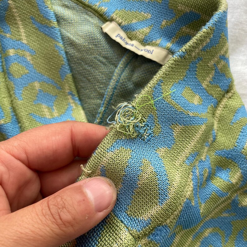 May include: A knit garment featuring a blue and green abstract pattern with a Paloma Wool brand label sewn into the inner collar. The fabric shows a visible area of loose, frayed threads and unraveling stitching along the top edge of the collar.