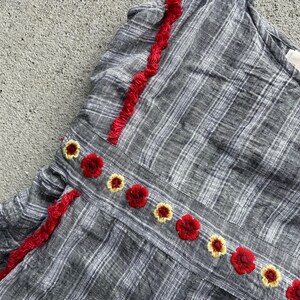 K&ouml;nnte beinhalten: Ein graues kariertes Oberteil mit roter Bord&uuml;re und gestickten Blumen. Das Oberteil hat eine Reihe von roten und gelben gestickten Blumen entlang der N&auml;hte. Die Bord&uuml;re ist leuchtend rot und verleiht dem Kleidungsst&uuml;ck einen Farbtupfer.