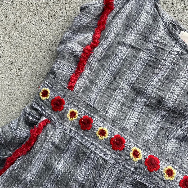 K&ouml;nnte beinhalten: Ein graues kariertes Oberteil mit roter Bord&uuml;re und gestickten Blumen. Das Oberteil hat eine Reihe von roten und gelben gestickten Blumen entlang der N&auml;hte. Die Bord&uuml;re ist leuchtend rot und verleiht dem Kleidungsst&uuml;ck einen Farbtupfer.