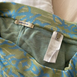 May include: A knit garment featuring a sage green and light blue abstract pattern. The interior includes a beige brand label reading paloma-wool and a white care tag marked with the size M. The fabric has a textured, woven appearance throughout.