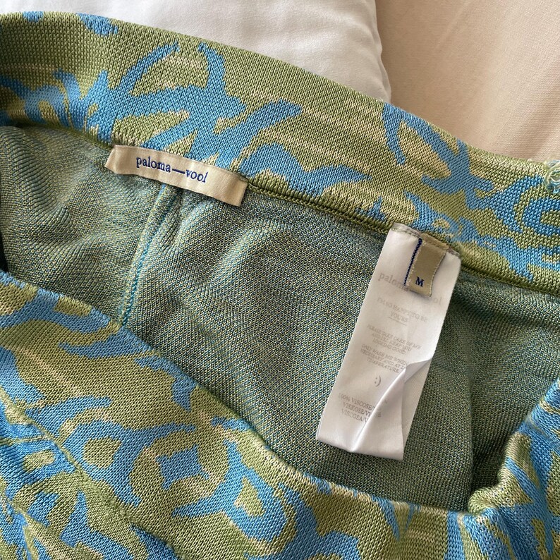 May include: A knit garment featuring a sage green and light blue abstract pattern. The interior includes a beige brand label reading paloma-wool and a white care tag marked with the size M. The fabric has a textured, woven appearance throughout.