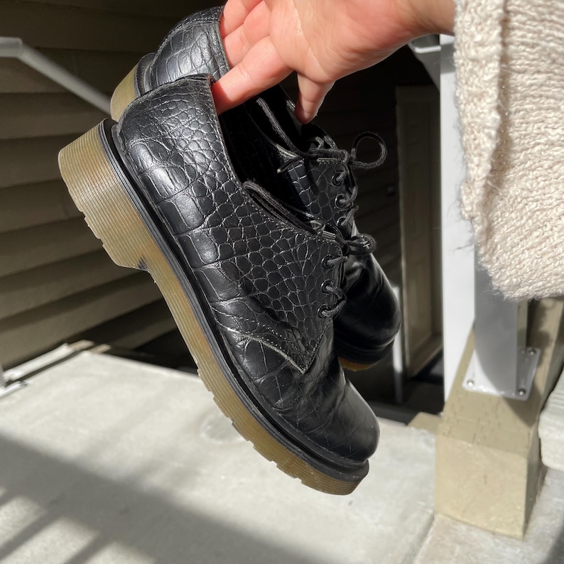 Puede incluir: Zapatos Oxford de cuero negro con textura de piel de cocodrilo sint&eacute;tica. Los zapatos tienen un dise&ntilde;o con cordones y una suela gruesa de color marr&oacute;n claro. Los zapatos est&aacute;n sostenidos por una mano.
