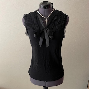 May include: A black sleeveless top featuring a V-neckline with a satin ribbon tie, ruffled trim along the shoulders, and a fitted silhouette. A white beaded necklace with a cross pendant is worn around the neck of the display mannequin.