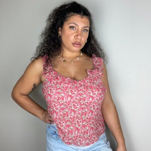 May include: A woman wearing a sleeveless, pink and white floral print top with a ruffled neckline. She is wearing a gold necklace with charms.