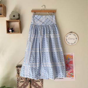 Pu&ograve; includere: Una gonna azzurra con un motivo geometrico di quadrati bianchi e piccoli motivi di ancore blu navy. La gonna &egrave; appesa a una gruccia di legno contro un muro neutro.