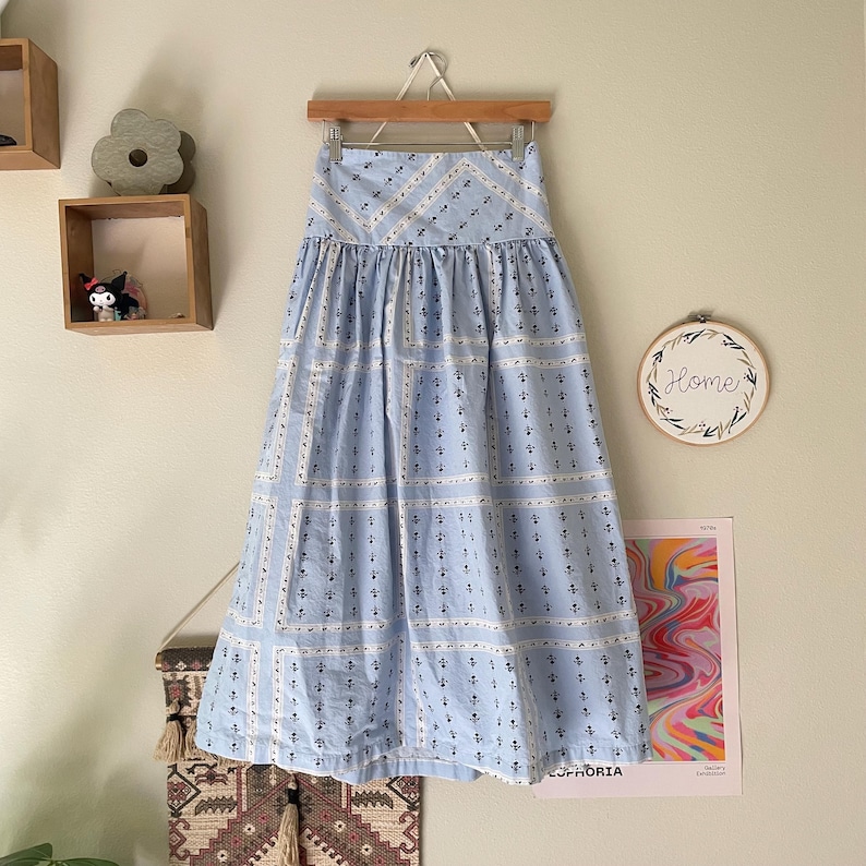 Pu&ograve; includere: Una gonna azzurra con un motivo geometrico di quadrati bianchi e piccoli motivi di ancore blu navy. La gonna &egrave; appesa a una gruccia di legno contro un muro neutro.