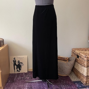 May include: A floor-length black skirt with a smooth, solid-colored fabric texture. The garment features a simple, straight-cut silhouette with an elasticized waistband, displayed on a mannequin torso against a neutral wall.