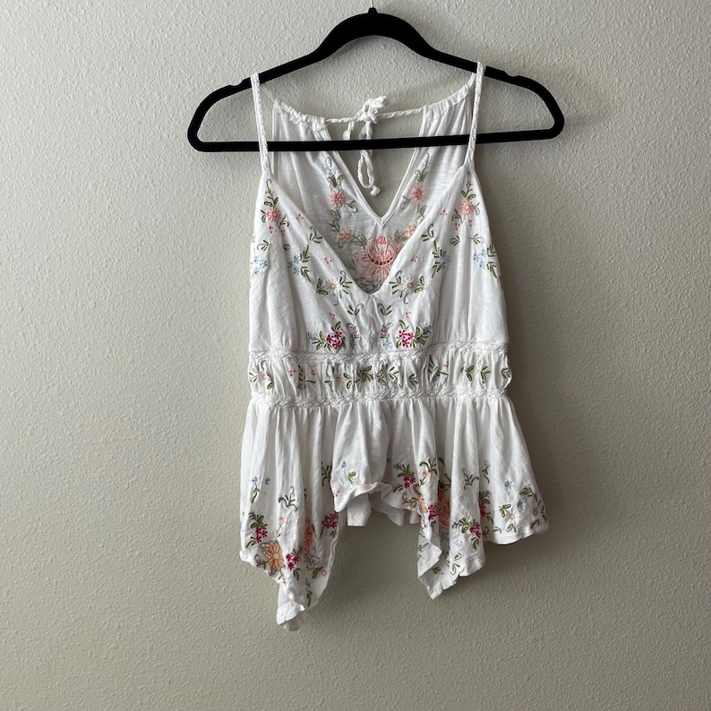 May include: White tank top with a V-neck and floral embroidery in pink, green, and orange. The top has a gathered waist and a flowing, asymmetrical hemline. It hangs on a black hanger against a neutral wall.