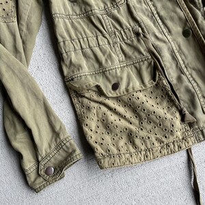Pode incluir: Casaco utilit&aacute;rio verde-oliva com detalhes de ilh&oacute;s nos bolsos e ombros. O casaco apresenta v&aacute;rios bolsos, cintura com cord&atilde;o e fechos de bot&atilde;o. As mangas t&ecirc;m punhos abotoados. O tecido tem um aspeto ligeiramente desgastado.
