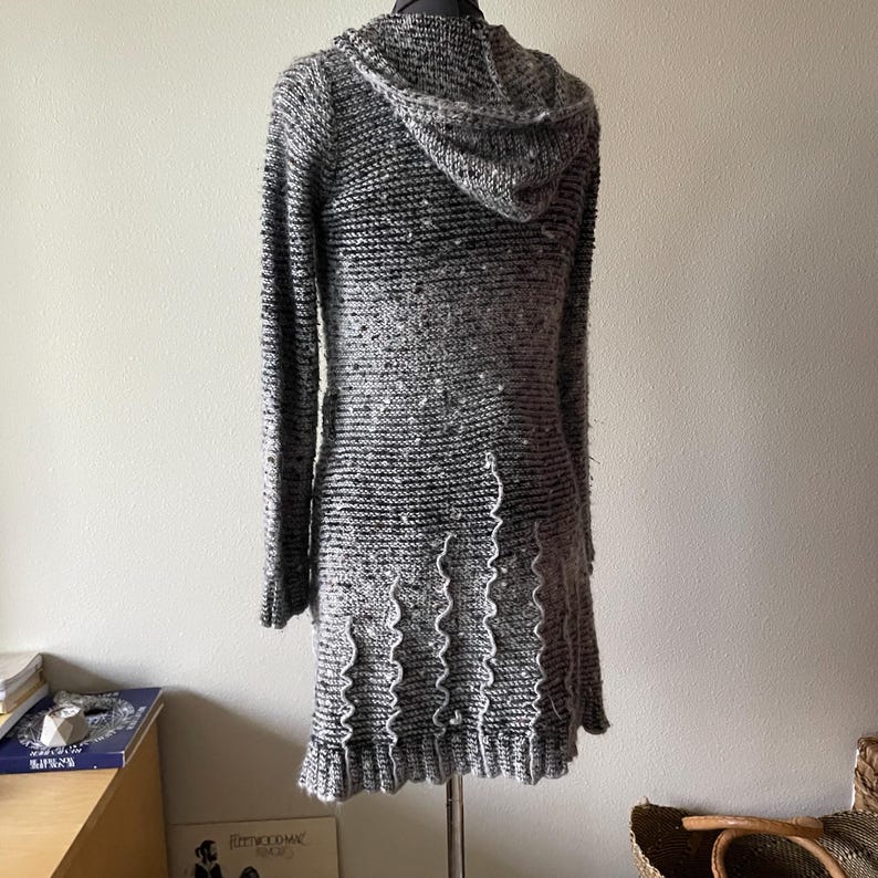 Puede incluir: Vestido de punto con capucha gris y negro con mangas largas, expuesto en un maniqu&iacute;. El vestido presenta un patr&oacute;n de punto texturizado, una capucha y un dobladillo con volantes. El vestido llega aproximadamente a la rodilla.