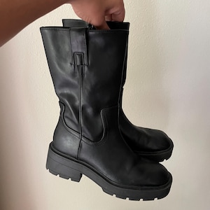 May include: A pair of black mid-calf boots with a chunky sole. The boots have a smooth, leather-like appearance and a small strap detail near the top. The soles are thick and provide a sturdy base. The boots are held up by a hand.