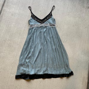 K&ouml;nnte beinhalten: Ein hellblaues Maxikleid mit schwarzer Spitzenborte am Ausschnitt und schwarzem Samtbesatz am Saum. Das Kleid hat ein figurbetontes Oberteil und einen flie&szlig;enden Rock.