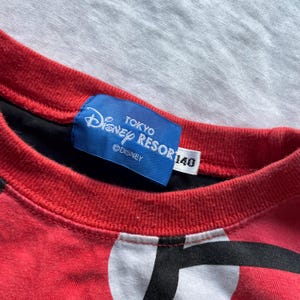 May include: Red and black crew neck shirt with a white and black graphic design. The shirt features a blue tag with "Disney Resort Tokyo" text and a size 140 label.