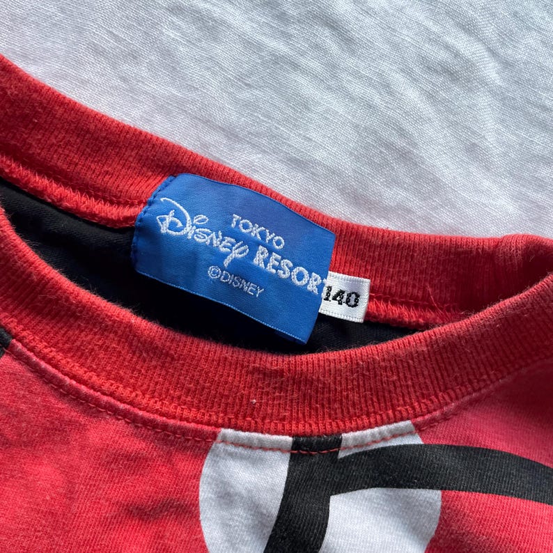 May include: Red and black crew neck shirt with a white and black graphic design. The shirt features a blue tag with "Disney Resort Tokyo" text and a size 140 label.