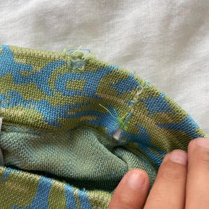 May include: A knit fabric garment featuring a repeating abstract pattern in shades of light blue and olive green. The top edge shows visible fraying and loose threads along the seam, with a small white tag partially tucked into the inner fold.