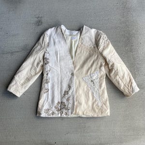 Op de afbeelding: Crèmekleurige blazer met een gesplitst ontwerp. Eén kant is gebroken wit met geborduurde bloempatronen, de andere is lichtcrème met kanten details. Voorzien van een enkele knoopsluiting en lange mouwen.