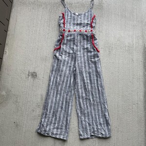 K&ouml;nnte beinhalten: Grau-wei&szlig; gestreifter Jumpsuit mit Spaghettitr&auml;gern. Der Jumpsuit hat rote Verzierungen an den R&auml;ndern des Oberteils und der Taschen sowie ein dekoratives Band aus roten und wei&szlig;en Kreisen an der Taille. Weite Hosenbeine sind sichtbar.