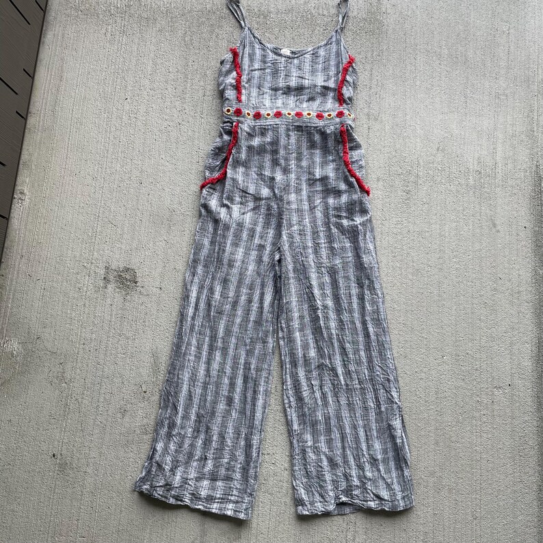 K&ouml;nnte beinhalten: Grau-wei&szlig; gestreifter Jumpsuit mit Spaghettitr&auml;gern. Der Jumpsuit hat rote Verzierungen an den R&auml;ndern des Oberteils und der Taschen sowie ein dekoratives Band aus roten und wei&szlig;en Kreisen an der Taille. Weite Hosenbeine sind sichtbar.