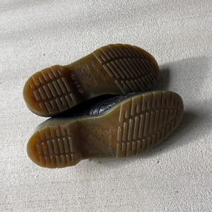 Puede incluir: Un par de zapatos de cuero negro con un patr&oacute;n texturizado. Las suelas marrones tienen un dibujo de la banda de rodadura y el nombre de la marca es visible. Los zapatos est&aacute;n sobre una superficie gris claro.