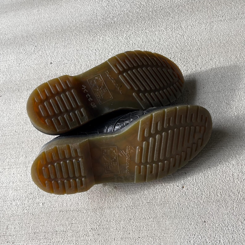 Puede incluir: Un par de zapatos de cuero negro con un patr&oacute;n texturizado. Las suelas marrones tienen un dibujo de la banda de rodadura y el nombre de la marca es visible. Los zapatos est&aacute;n sobre una superficie gris claro.