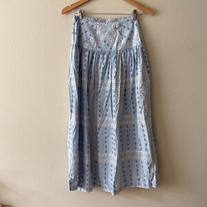 Pu&ograve; includere: Gonna azzurra con un motivo ripetuto di disegni floreali e geometrici bianchi e blu navy. La gonna &egrave; appesa a una gruccia di legno contro un muro neutro.