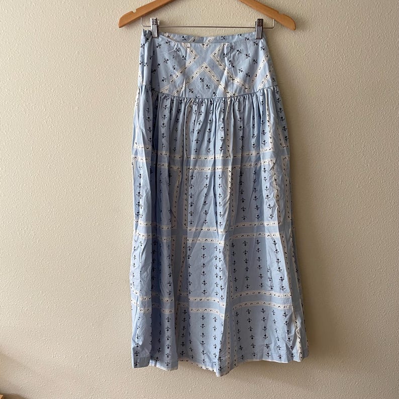 Pu&ograve; includere: Gonna azzurra con un motivo ripetuto di disegni floreali e geometrici bianchi e blu navy. La gonna &egrave; appesa a una gruccia di legno contro un muro neutro.