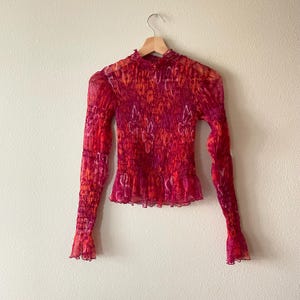 May include: A long-sleeved, sheer top in shades of red, orange, and pink, hanging on a wooden hanger. The top features a high neckline, ruffled hem, and a textured, smocked bodice. The sleeves have a flared, ruffled cuff.
