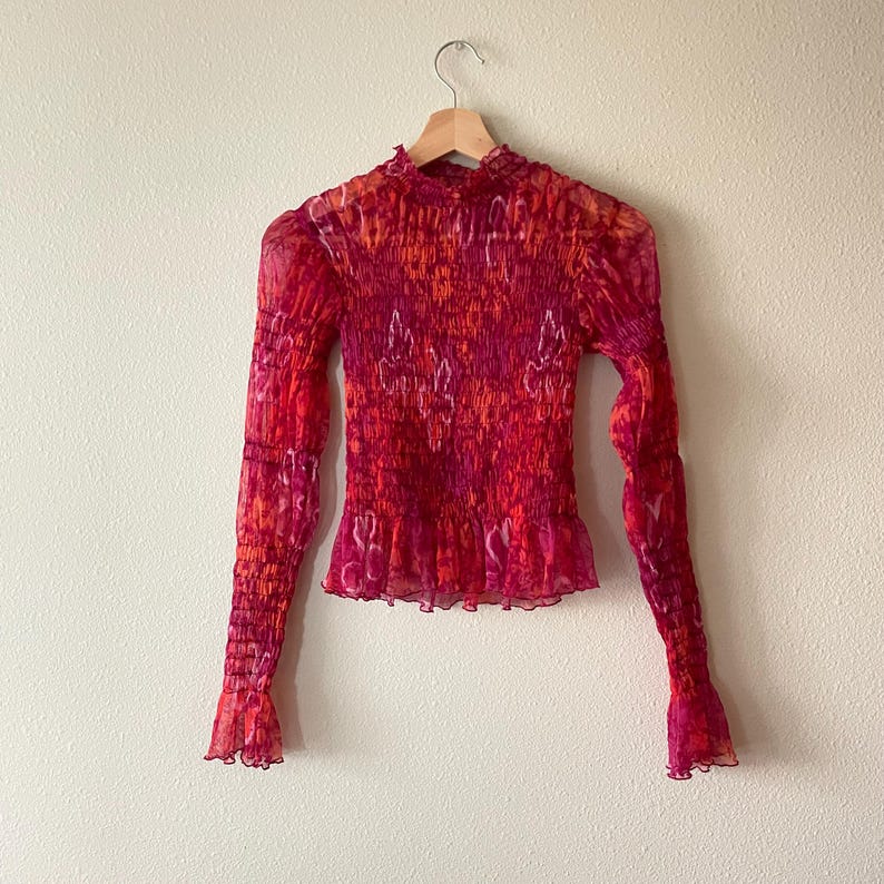May include: A long-sleeved, sheer top in shades of red, orange, and pink, hanging on a wooden hanger. The top features a high neckline, ruffled hem, and a textured, smocked bodice. The sleeves have a flared, ruffled cuff.