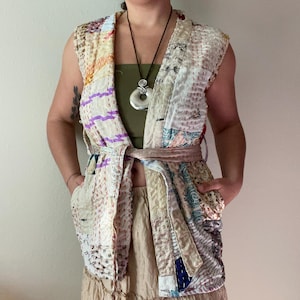 May include: A sleeveless, patchwork vest in cream, beige, and pastel colors. The vest features a wrap-around tie and pockets. The garment is made of various fabric scraps sewn together, creating a unique, textured look. The vest is worn over a green top and tan skirt.