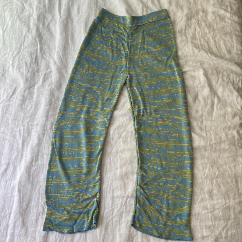 May include: Full-length pants featuring a high-waisted design and a wide-leg silhouette. The fabric displays a horizontal, abstract pattern in alternating shades of light blue and olive green, creating a textured, layered visual effect throughout.