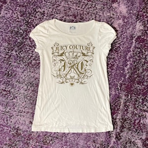 Pode incluir: Uma t-shirt branca de manga curta com decote redondo e mangas franzidas. A parte frontal apresenta uma impressão gráfica em dourado metálico com o texto Juicy Couture, uma coroa, âncoras e andorinhas centradas na zona do peito.
