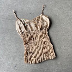 Pode incluir: Uma blusa de cetim bege com saia plissada e bordados florais. A blusa tem alças finas e um decote redondo.