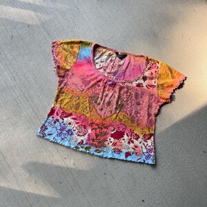 Pode incluir: Uma camisa de manga curta colorida, estilo tie-dye, com decote redondo e um design de contas. A camisa tem um padrão floral com cores rosa, azul, amarelo e verde.