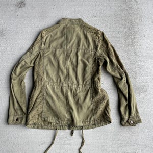 Pode incluir: Casaco verde-oliva com gola alta, mangas compridas e cintura com cord&atilde;o. O casaco apresenta detalhes decorativos vazados nos ombros e laterais. Possui fecho de correr e punhos abotoados.