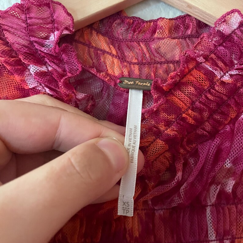 May include: Close-up of a sheer, ruffled top in shades of pink and orange. The top has a textured, layered appearance. A white tag with the brand "Free People" is visible, along with the size "XS TP".