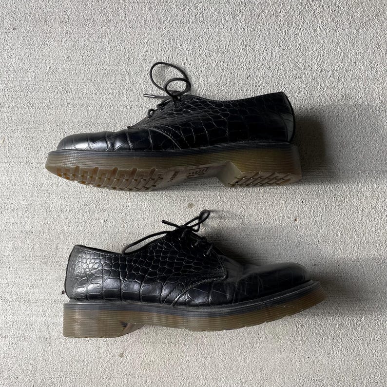 Puede incluir: Par de zapatos Oxford de cuero negro con textura de piel de cocodrilo. Los zapatos tienen cierre de cordones y una suela gruesa de color marr&oacute;n claro. Los zapatos se muestran sobre una superficie de hormig&oacute;n gris.