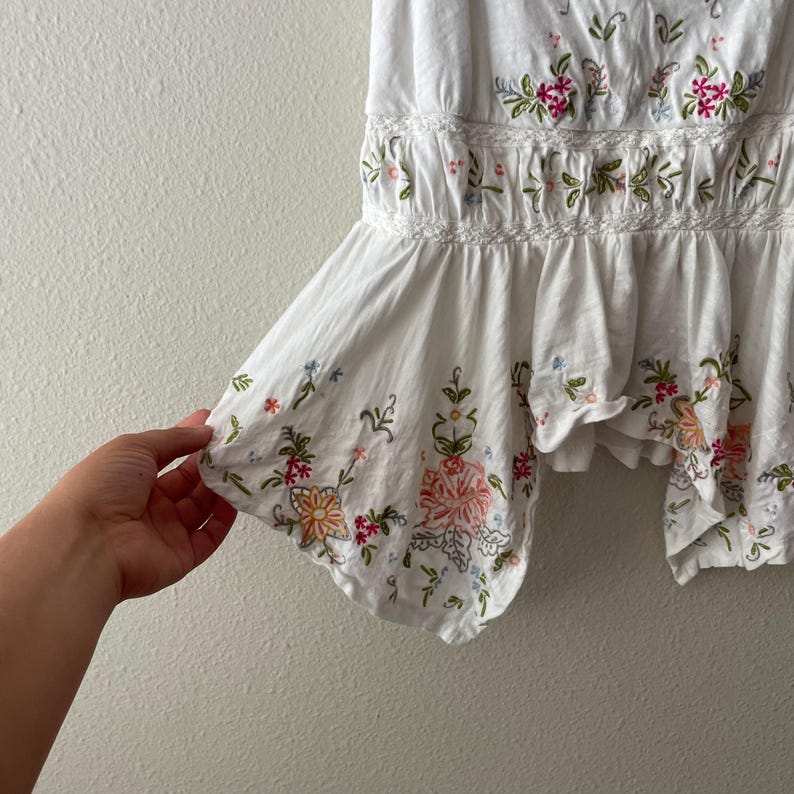 May include: White top with floral embroidery in pink, orange, and green. The top features a ruffled hem and delicate detailing around the waist. The embroidery includes various floral and leaf patterns.