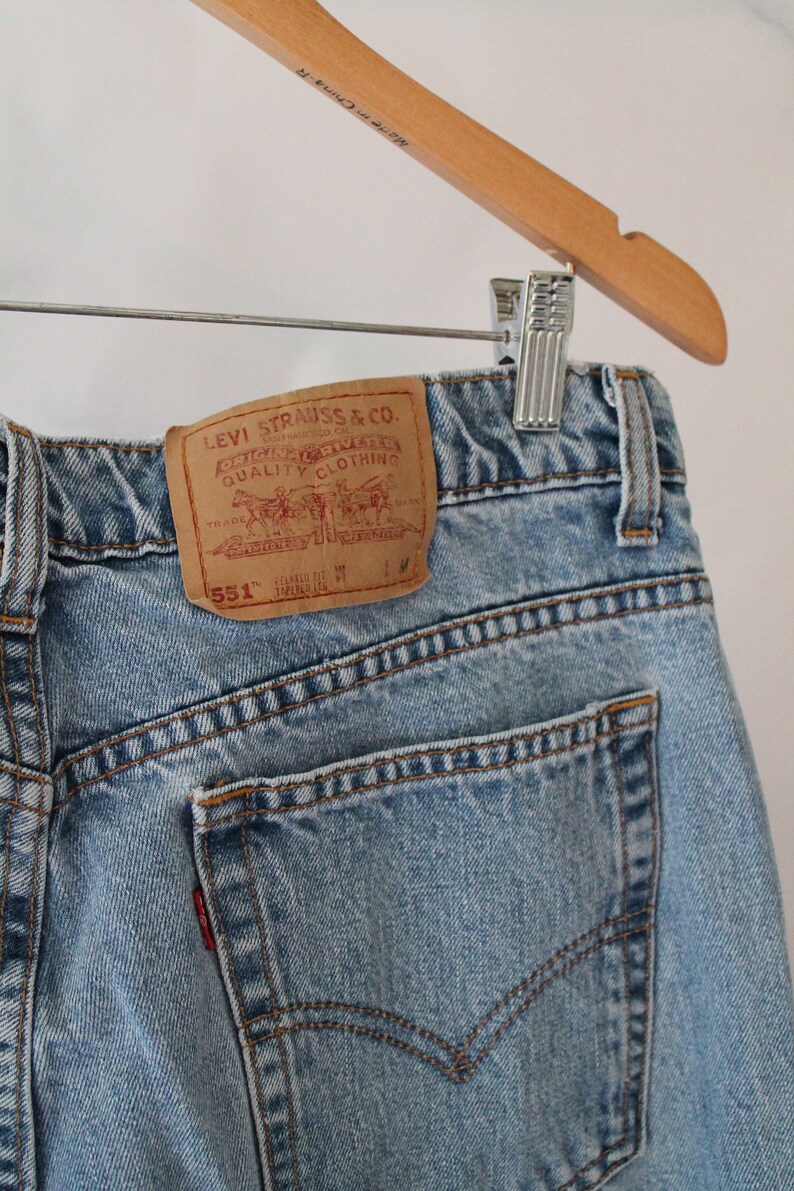 Puede incluir: Un par de pantalones vaqueros de mezclilla azul claro con un parche de cuero marr&oacute;n que dice "Levi Strauss & Co. Original Riveted Quality Clothing" y "551" en texto blanco.