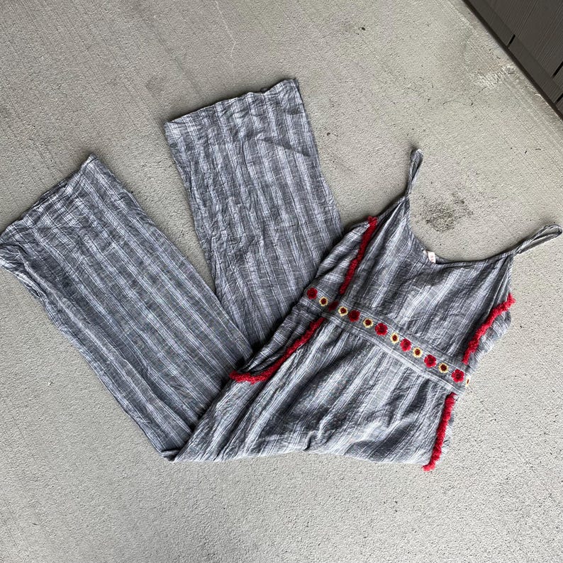 K&ouml;nnte beinhalten: Grau-wei&szlig; gestreifter Jumpsuit mit Spaghettitr&auml;gern. Das Oberteil hat rote Verzierungen und dekorative goldfarbene &Ouml;sen. Der Jumpsuit hat weite Beine und ist aus leichtem Stoff gefertigt.